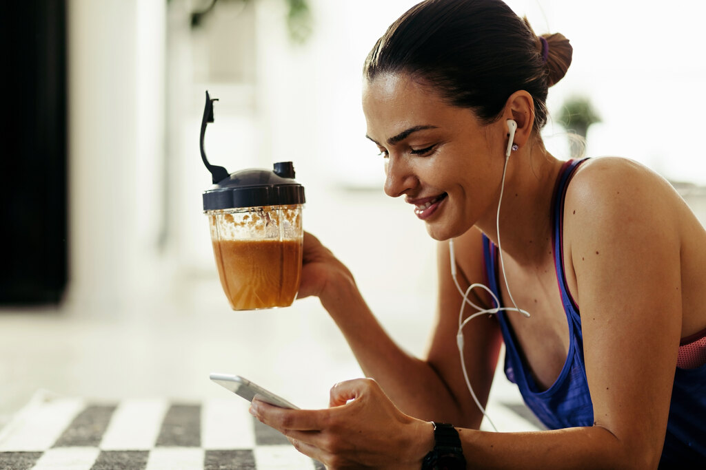 Mulher sorridente olhando para o celular enquanto segura uma bebida na outra mão