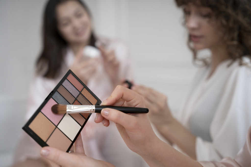Mãos de uma maquiadora segurando um pincel de maquiagem em primeiro plano, com jovem ao fundo aplicando creme