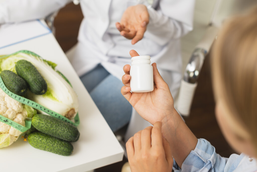 Mãos de uma mulher caucasiana segurando um frasco de capsulas com médico ao fundo explicando