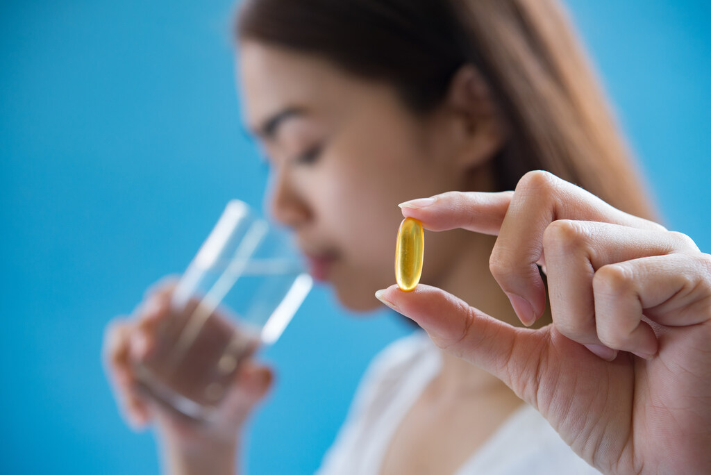 Mulher caucasiana tomando água no segundo plano enquanto segura uma capsula de medicamento em primeiro plano
